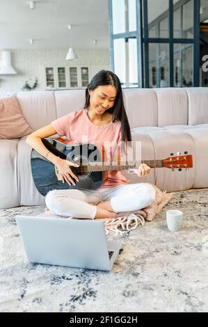 Vertical image of pretty Asian woman with long black hair playing guitar, sitting on the floor in living room, giving online music classes, teaching, spending weeked with online friends from the laptop Stock Photo