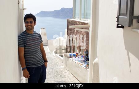 A Portrait of a young brown asian Indian ethnicity man with stubble and sunglasses smiling with a hand in the pocket against beautiful sea scape of Gr Stock Photo