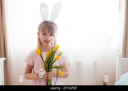 Easter concept. Small smiling girl in white fluffy bunny ears and pink dress holding yellow tulip flowers and colorful egg. Room interior. High qualit Stock Photo