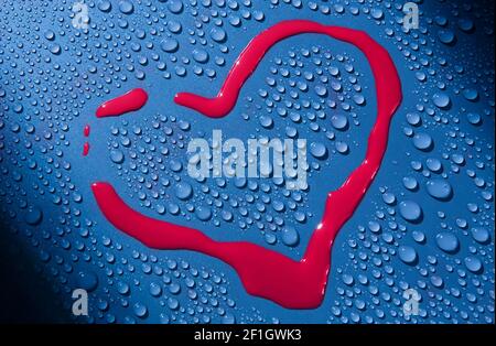 Drawing in red paint in shape of heart on glass surface surrounded by water droplets. Declaration of love. Closeup. Background. Stock Photo