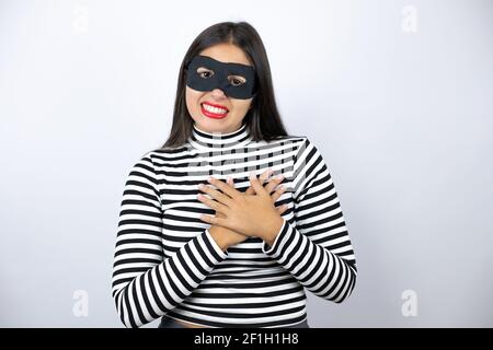 Young beautiful brunette burglar woman wearing mask smiling with her hands on her chest and grateful gesture on her face. Stock Photo