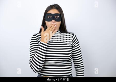 Young beautiful brunette burglar woman wearing mask covering mouth with hand, shocked and afraid for mistake. surprised expression Stock Photo