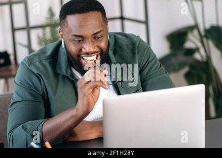 Satisfied happy black male freelancer, programmer, sits at his desk, holding his eyeglasses in hands, looks at a laptop, received an great news or message,saw a financial statement,good profit or deal Stock Photo