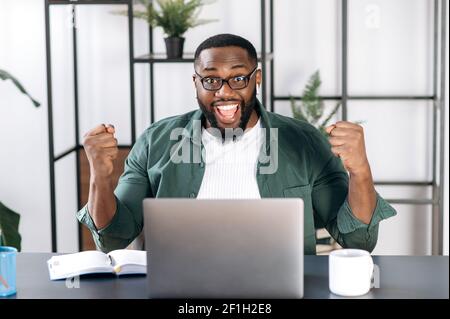 Happy African American male freelancer dressed in stylish clothes, sitting at his work desk using laptop, rejoicing in success, win or good profit, looking at the camera and gesturing with his hands Stock Photo