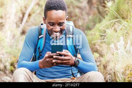 Young African ethnicity man video chatting via cellphone with somebody and cheerfully laughing as he having a hiking walk in the forest. Happy people, Stock Photo