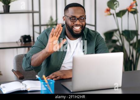 Online video communication. Happy smiling African American guy, freelancer or student, waving hand, greeting colleague, uses laptop for video conference with coworkers, telecommunication concept Stock Photo