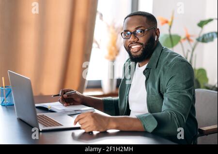 Attractive friendly African American guy, successful freelancer, manager or student, sits at his desk at home or office, uses a laptop for work, looks at the camera and smiles Stock Photo