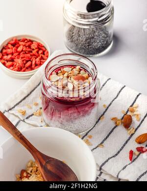 Cooking healthy breakfast - coconut milk chia pudding, goji berries, flax, pumpkin seeds, nuts and granola ingredients. Table set with striped napkin Stock Photo