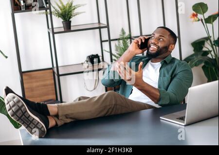 Relaxed confident african american male freelancer or business man sitting at work desk in the office with legs on table and talking on the cellphone with business partner, gesturing hand, smiling Stock Photo