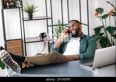 Relaxed confident satisfied multiethnic black male freelancer or modern business man sitting at work desk in the office with legs on table and talking on the cellphone with business partner, smiling Stock Photo
