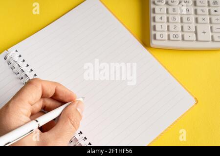 close up on hand holding pencil writing on notebook by the calculator on yellow background - office and business or goals concept Stock Photo