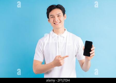 Smiling young man holds a modern smartphone in his hand and shows his finger on a blank white screen, isolated on a blue background. Stock Photo