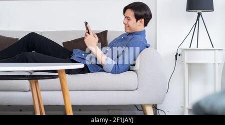 Young Asian man lying on sofa and using phone Stock Photo