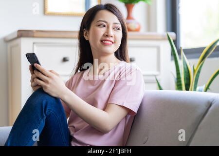 Beautiful young Asian woman sitting on sofa using her phone and looking out the window