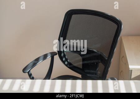 A black mesh-backed office armchair facing a beige-painted wall behind a table next to a wooden nightstand, sunlight streaming through the blinds onto Stock Photo