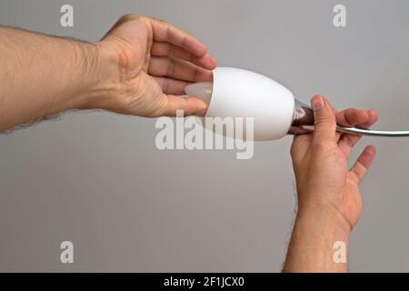 The left man's hand holds the arm of the chandelier the right hand screws or unscrews energy-saving candle-type lamp on white ceiling background. Conc Stock Photo