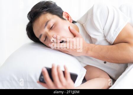 The young man is lying on the bed yawning, still holding the phone Stock Photo