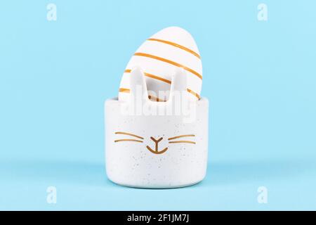 White Easter egg painted with golden stripes in cute bunny shaped egg cup on blue background Stock Photo