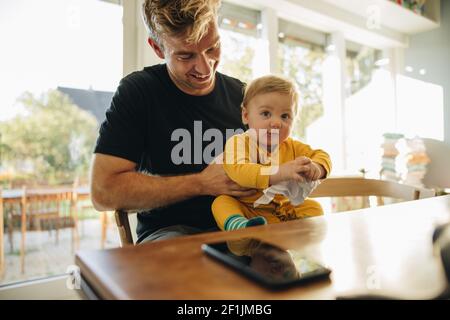 Smiling man with his son sitting at table. Father with his cute baby boy at home. Stock Photo