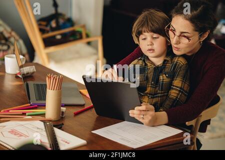 Woman with son sitting at home using digital tablet. Mother teaching son drawing on digital tablet. Stock Photo