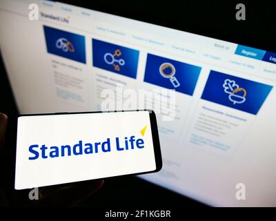 Person holding mobile phone with business logo of British insurance brand Standard Life on screen in front of web page. Focus on phone display. Stock Photo