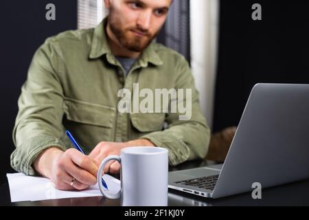 Working from home, casual business man using laptop computer, writing on notebook with cup of tea on table. Student reading e-book via computer Stock Photo