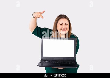 Freelancer. Caucasian smiling woman holds a laptop, pointing at the mock up screen with index finger. Plus size model. White empty blank pc. Concept Stock Photo