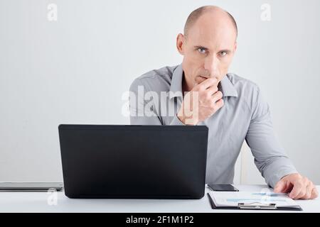 Portrait of pensive serious entrepreneur sitting at office desk with laptop and report and looking at camera Stock Photo