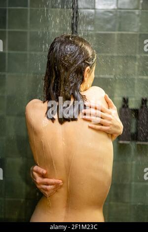 Woman taking shower and relaxing under warm running water in green bathroom. Hugs herself. Enjoy during hot water bath. Stock Photo