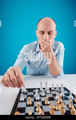 Frowning pensive mature man making move with chess piece,isolated on blue Stock Photo
