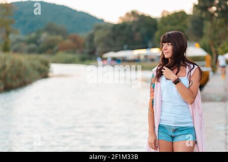 Beautiful woman with a tattoo on her arm posing near a lake. Embankment in the background. Summertime and vacation. Stock Photo