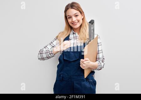 smiling female constructor holding level tool and paper clipboard in hands, posing at camera smiling. portrait of positive engineer in blue uniform Stock Photo