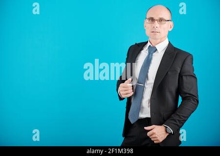 Portrait of confident mature businessman in glasses standing against blue background and looking at camera Stock Photo