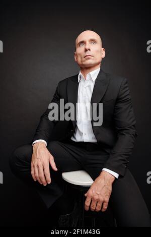 Portrait of confident elegant businessman in black suit sitting on stool and looking away Stock Photo