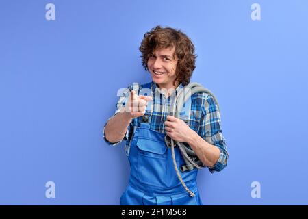 Happy male architect and plumber pointing finger at camera and smiling on blue background, portrait of young caucasian guy with curly hair dressed in Stock Photo