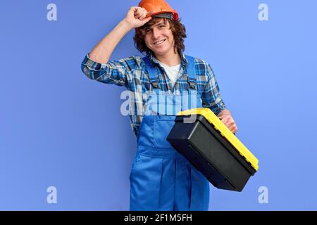 hard working engineer holding tool box in hands, having working day, wearing orange hardhat helmet and blue uniform, posing isolated over purple backg Stock Photo