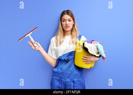 sad female with cleaning supplies stand dissatisfied by dirt in room she has to clean, isolated over purple background. portrait of maid in coveralls Stock Photo