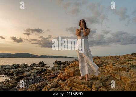 Pensive Woman in long white dress on with folded hands in front of face and closed eyes contemplating the sunset over the sea. Concept of experiencing Stock Photo