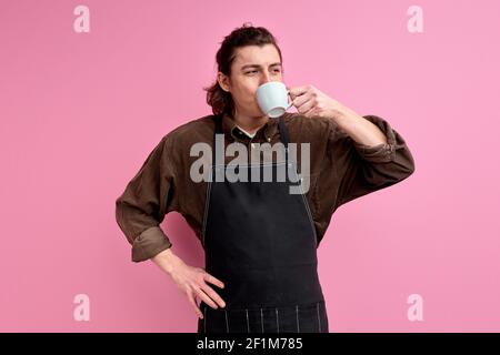 Young barista enjoying coffee in mug, wearing black apron. Handsome caucasian guy stand drinking beverage, isolated over pink studio background