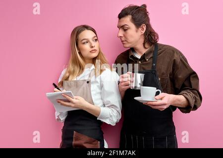 two young waiters in apron discuss orders, ready to serve clients, friendly waitress stand writing making notes and talking with colleague. isolated p Stock Photo
