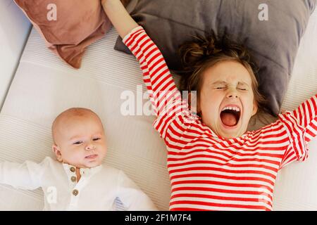 Girl lying with baby brother on bed and yawning Stock Photo