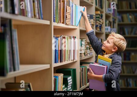 school boy taking books from shelves in library, with a stack of books in hands. child brain development, learn to read, cognitive skills concept Stock Photo