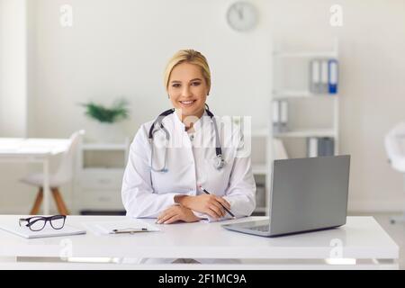 Friendly young woman, working as doctor, sitting at desk in hospital office, smiling and looking at camera. Stock Photo