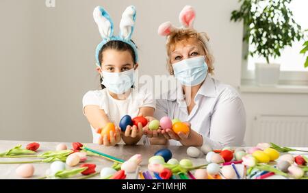 grandmother and granddaughter paint Easter eggs in the kitchen with tassels, phone, tulips and rabbits at home on quarantine with medical mask Stock Photo