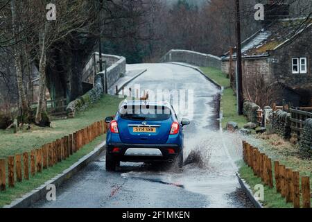 Blue Vauxhall Mokka car driving on country lane through puddle (splashing spraying water) on wet rainy day - Bolton Bridge, Yorkshire, England, UK. Stock Photo