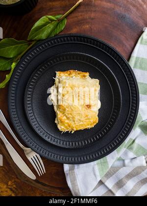 Delicious lasagna four cheeses on black ceramic plates, green leaves, cutlery and dishcloth with wooden base. Close-up, top view. Stock Photo