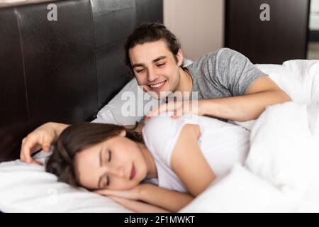Happy handsome young man lying and waking his woman in bed Stock Photo