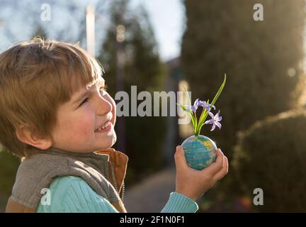 cute toddler boy holds a little ball - a globe from which small blue spring flowers grow. Eco concept, environmental education. Mother earth day. Sunn Stock Photo