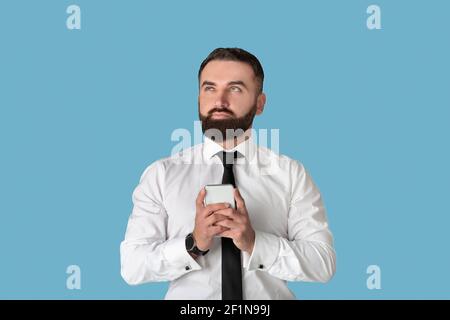 Pensive millennial office employee using smartphone for online work or communication against blue studio background. Thoughtful young businessman with Stock Photo