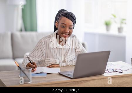 Young black lady having online training from home, using laptop and taking notes, copy space. Smiling african american woman attending online conferen Stock Photo
